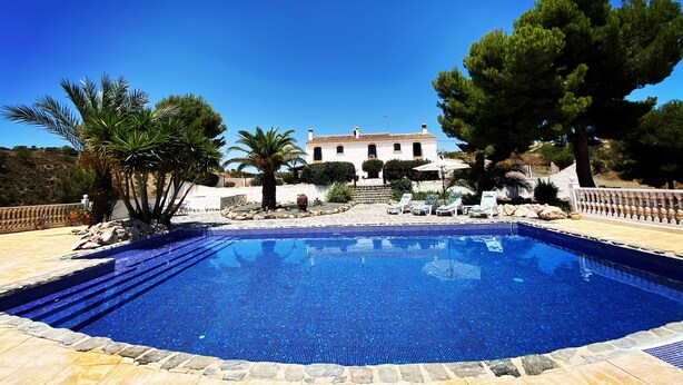 A swimming pool with a tiled edge, surrounded by a stone deck, palm trees, and a white building in the background. - Hotel Los Sibileys (Lorca)
