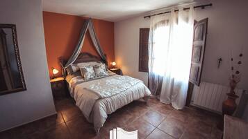 Un dormitorio con cama con dosel, dos lámparas de noche, un ventanal con cortinas traslúcidas y un radiador.