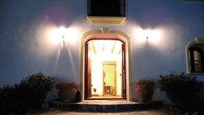A stone-paved entrance to a building with a lit doorway, flanked by two large potted plants. - Hotel Los Sibileys (Lorca)