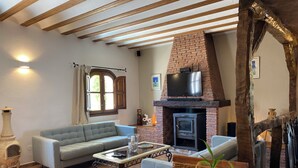 A living room with a brick fireplace, a sofa, a television, and a large window. - Hotel Los Sibileys (Lorca)