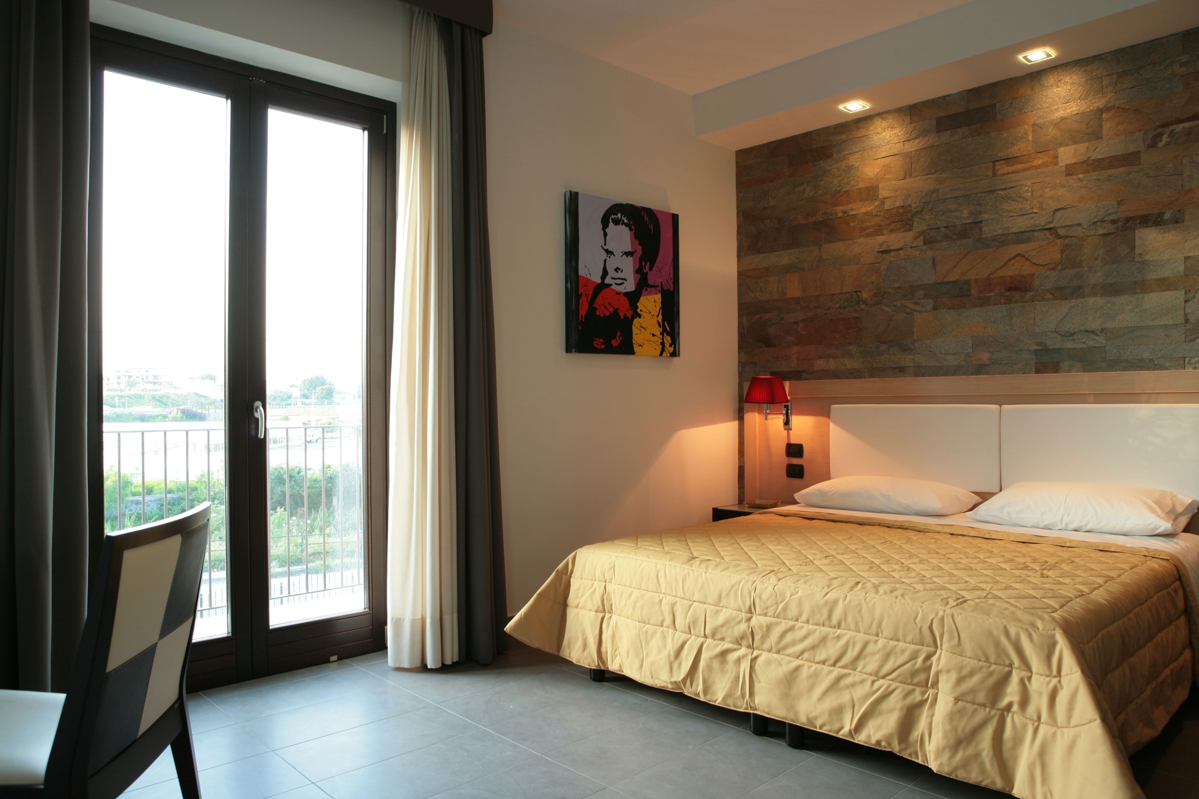 A hotel room with a bed, a chair, a painting, a nightstand, and a view through a sliding door.