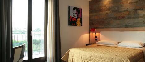 A hotel room with a bed, a chair, a painting, a nightstand, and a view through a sliding door.