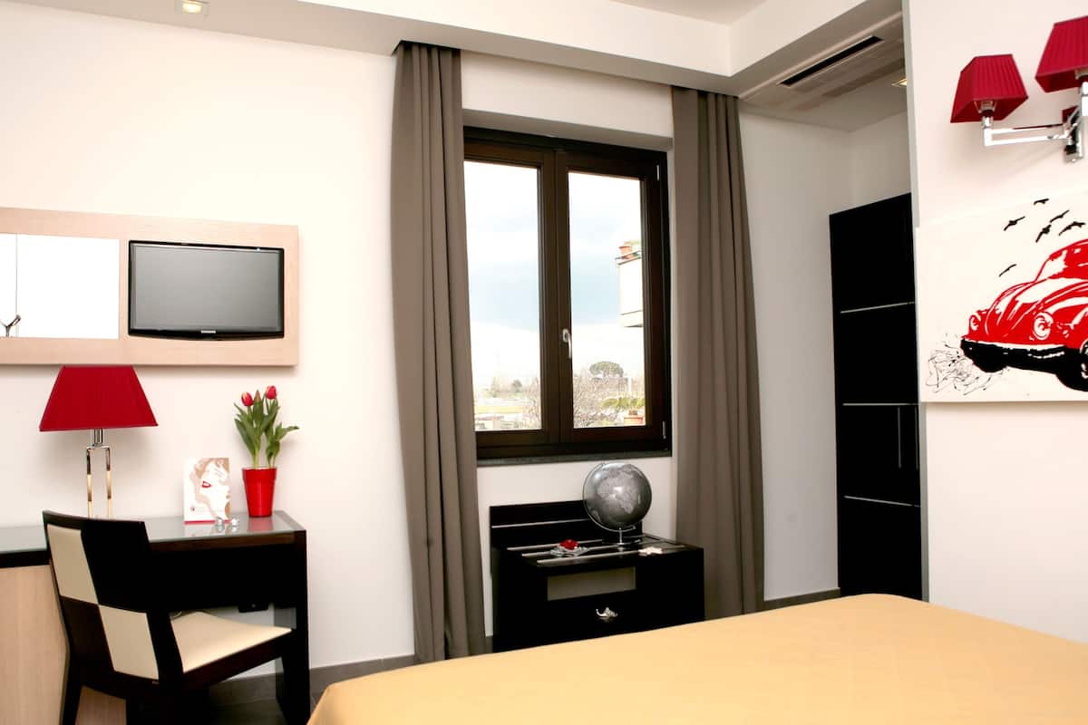 A hotel room with a bed, a desk with a lamp, a TV, a window with curtains, a red car painting, and a potted plant.