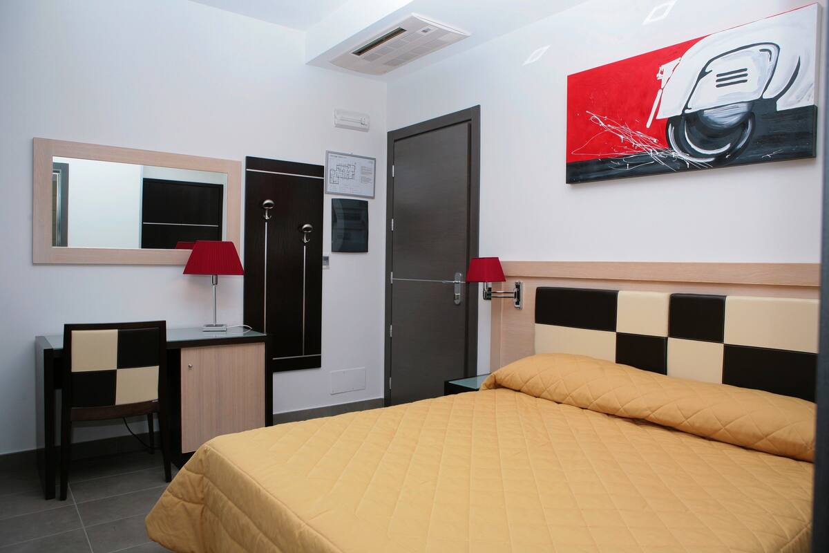 A hotel room with a bed, desk, chair, and a painting on the wall.