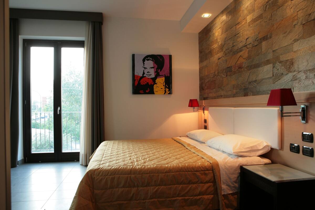 A hotel room with a bed, a red lamp, a painting on the wall, and a view through the sliding doors.