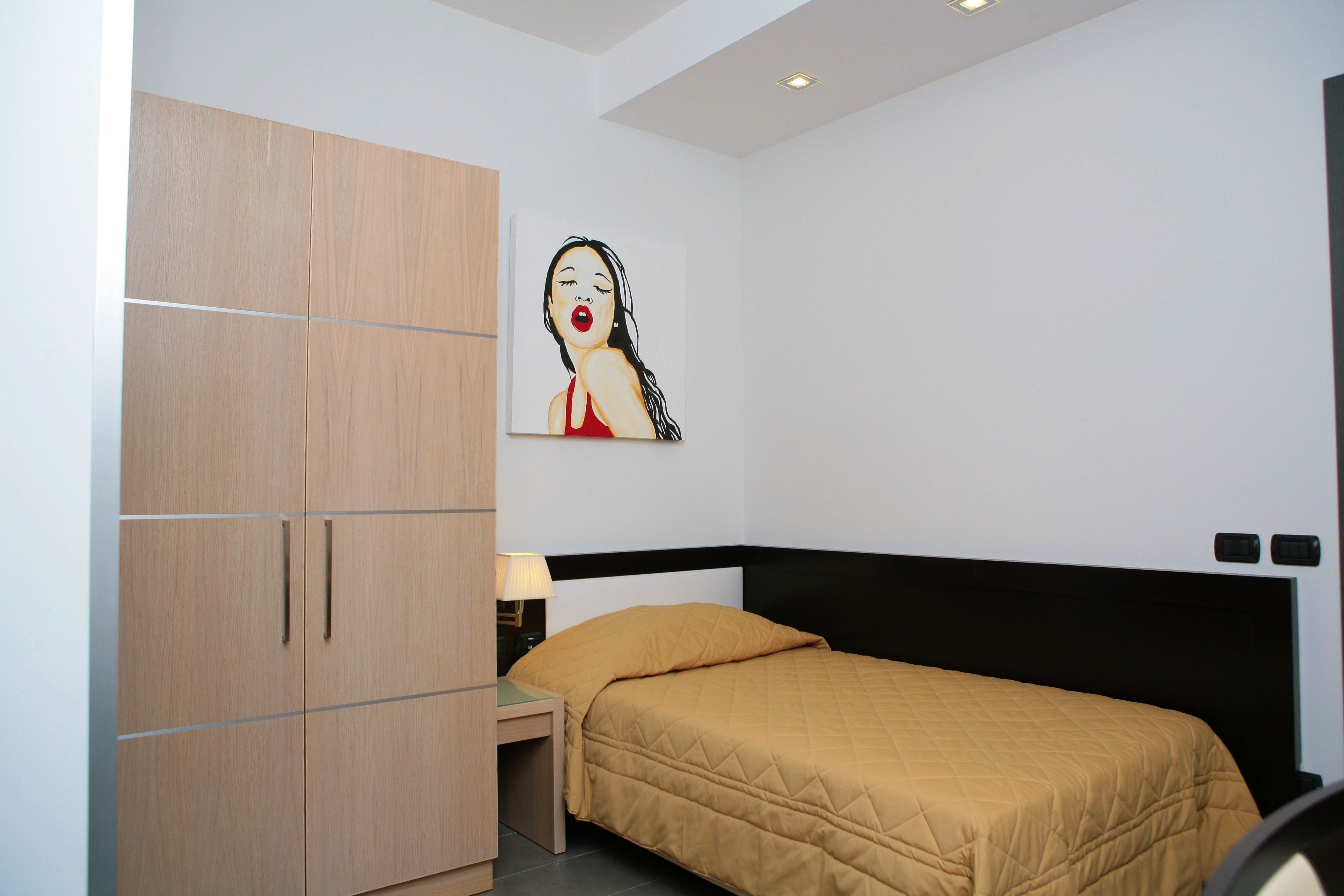 A bedroom with a bed, a wooden wardrobe, a nightstand, a lamp, and a wall art piece.