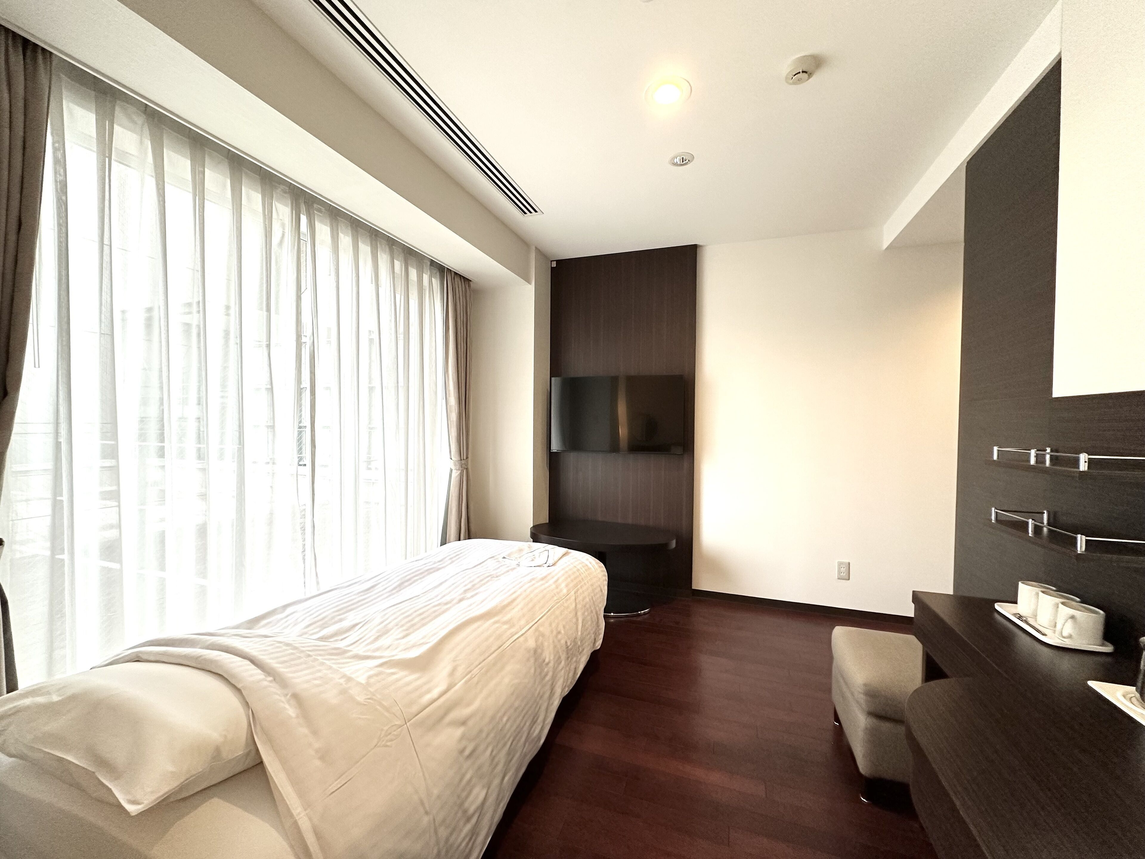 Une chambre d’hôtel moderne dotée d’un grand lit, d’un téléviseur à écran plat fixé au mur, d’un bureau avec une tasse et une soucoupe, et d’une fenêtre ornée de rideaux légers.
