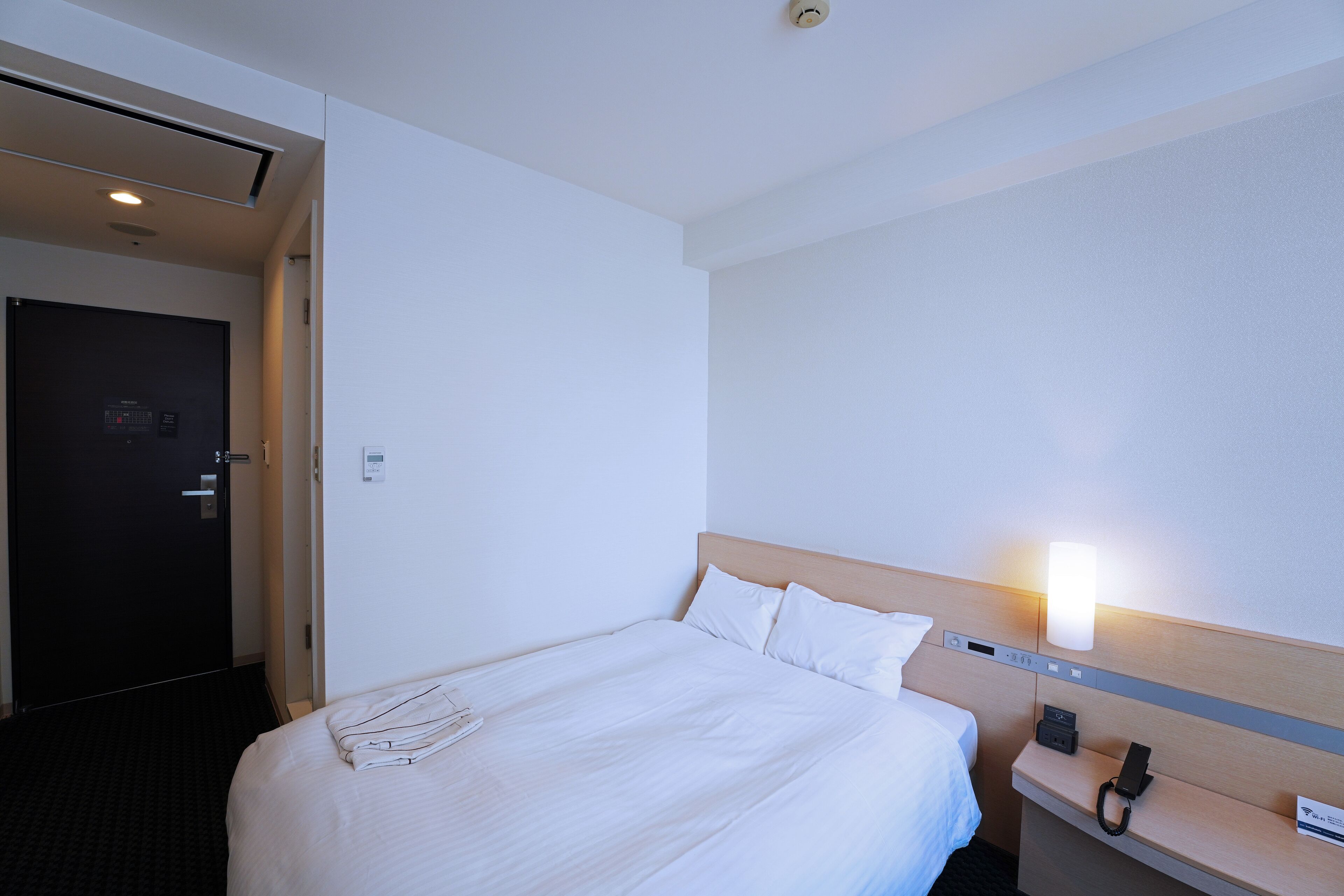 A hotel room with a neatly made bed, a bedside table with a lamp, and a door leading to another room.