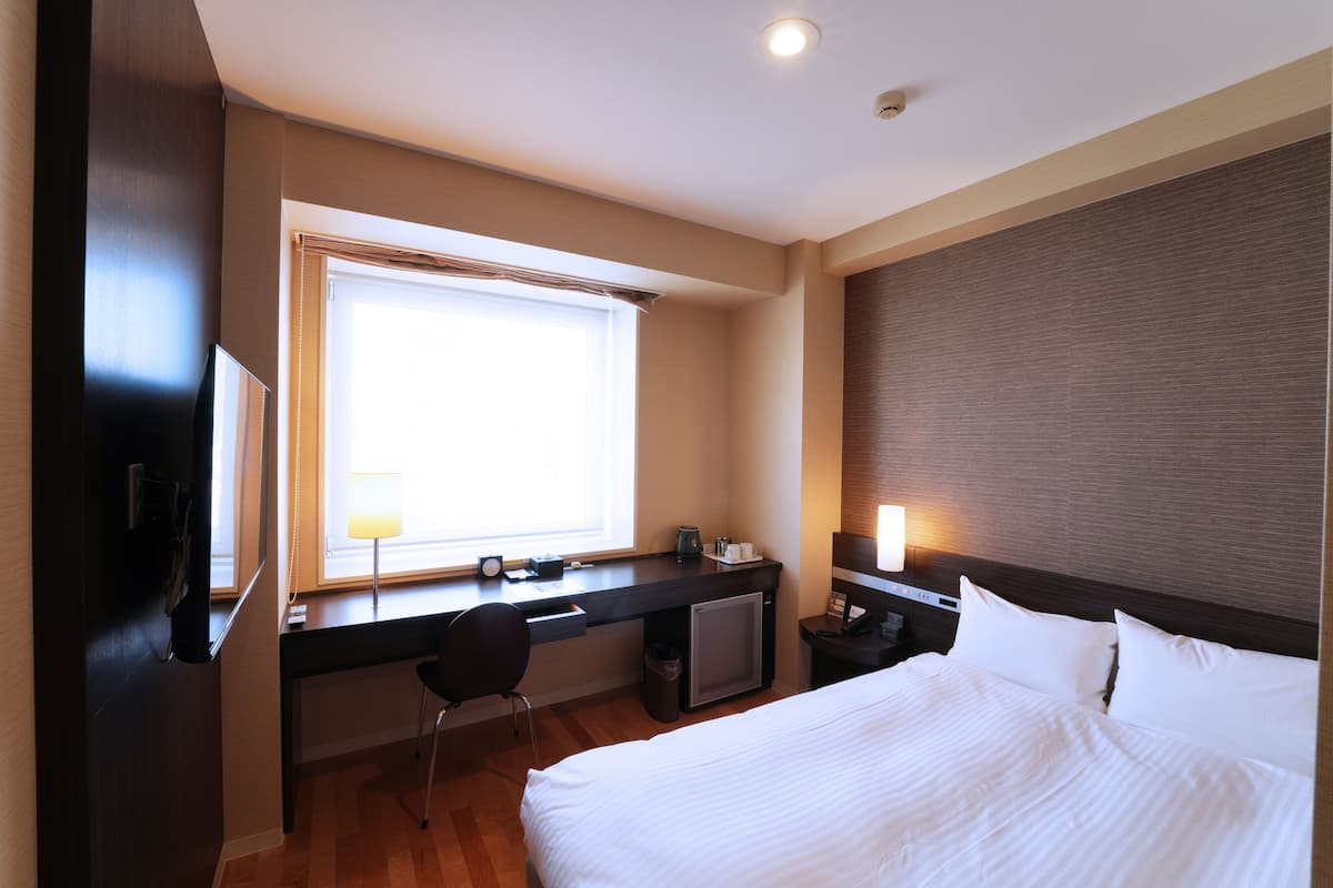 Quarto de hotel com cama, escrivaninha, cadeira e um pequeno frigorífico.