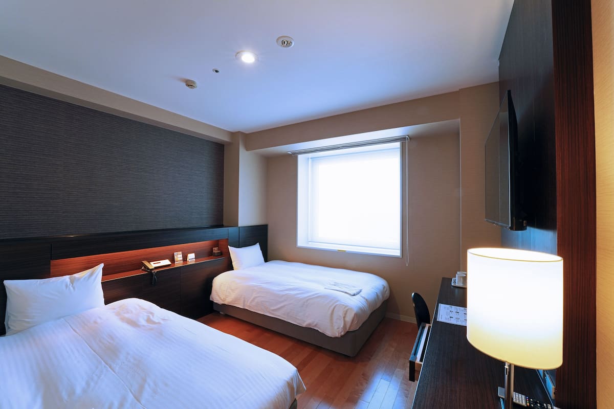 Quarto de hotel com duas camas, uma escrivaninha, luminária, televisão e janela com persianas.
