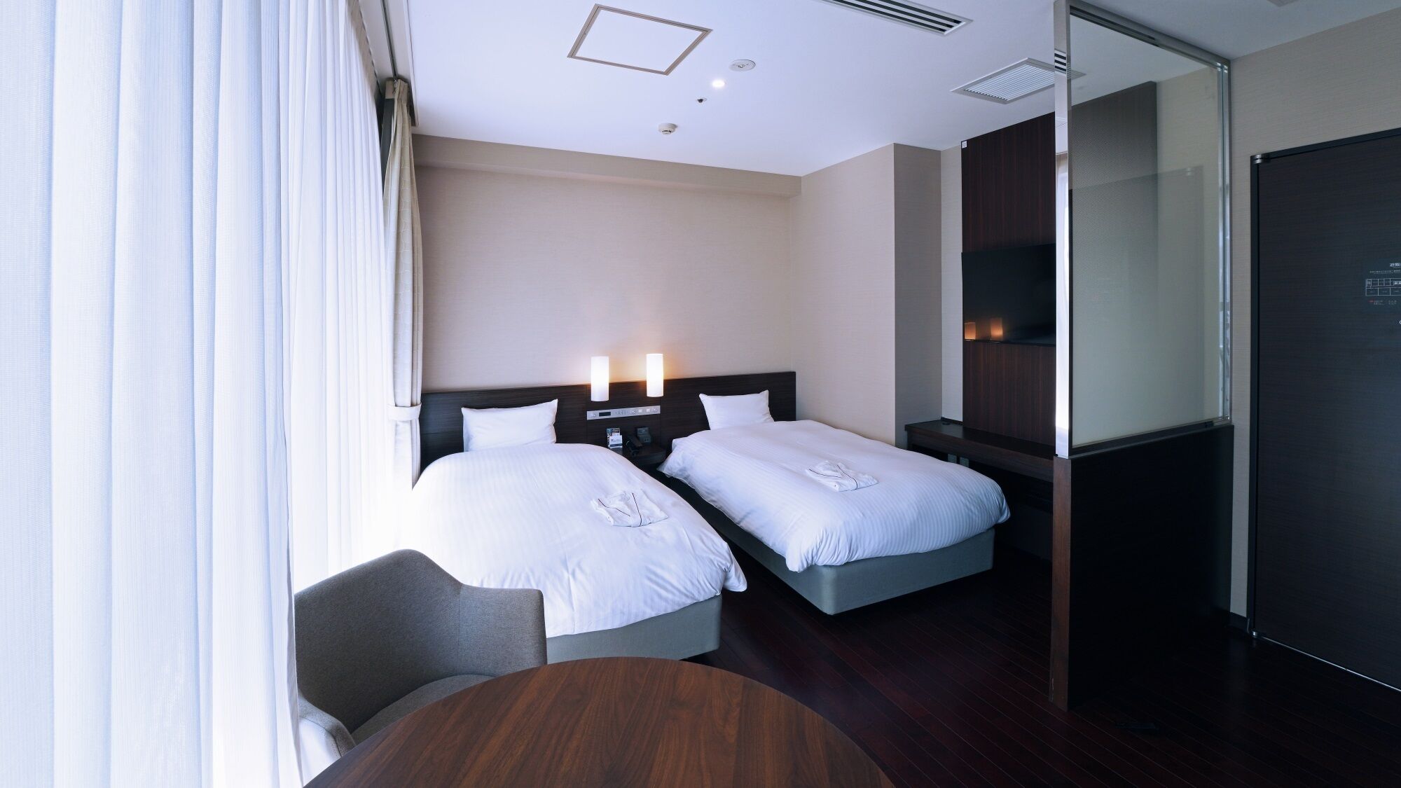 A hotel room with two beds, a desk, a chair, a TV, and a glass partition.