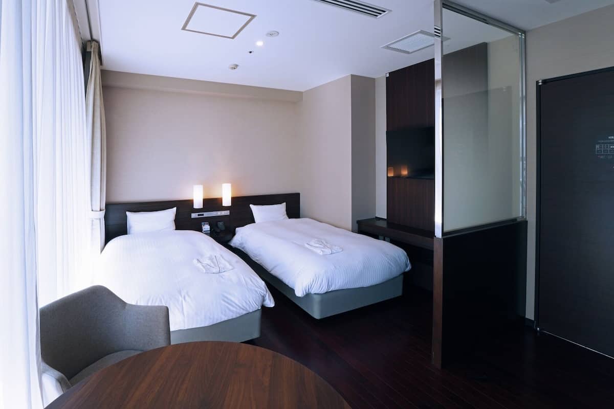 Quarto de hotel com duas camas, uma escrivaninha, uma cadeira, televisão e um painel de vidro.