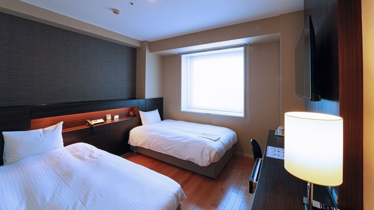 A hotel room with two beds, a desk with a lamp, a TV, and a window.