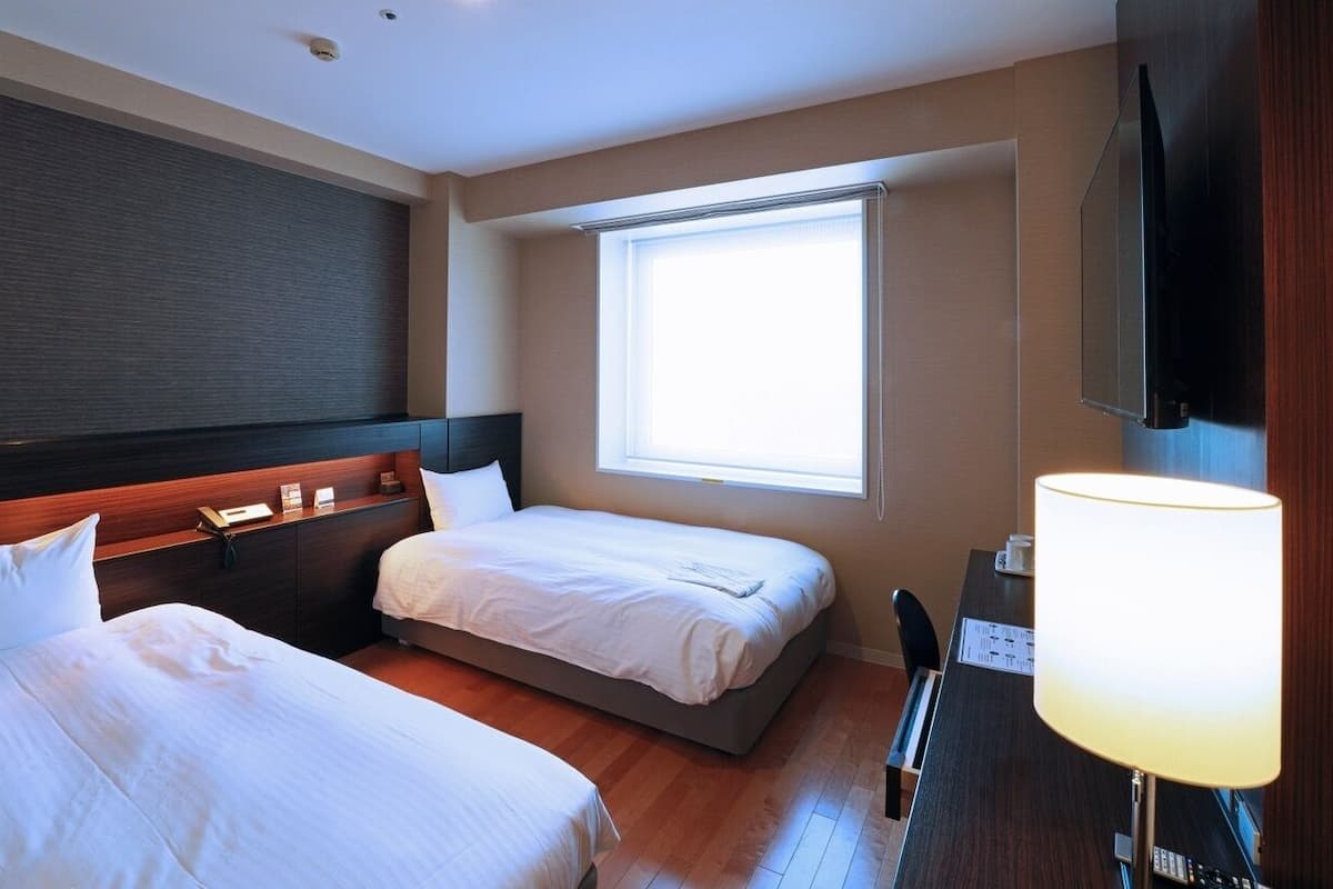 Quarto de hotel com duas camas, uma secretária com luminária, televisão e janela.