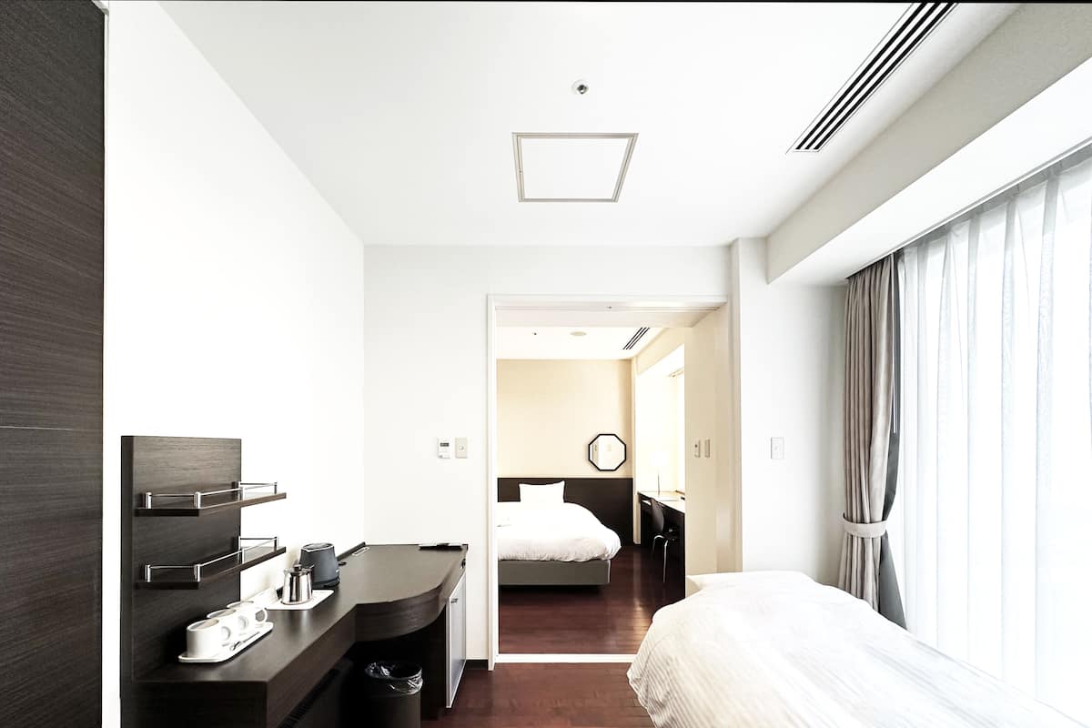 Quarto de hotel moderno com cama, escrivaninha com cadeira, lixeira pequena e janela com cortinas.