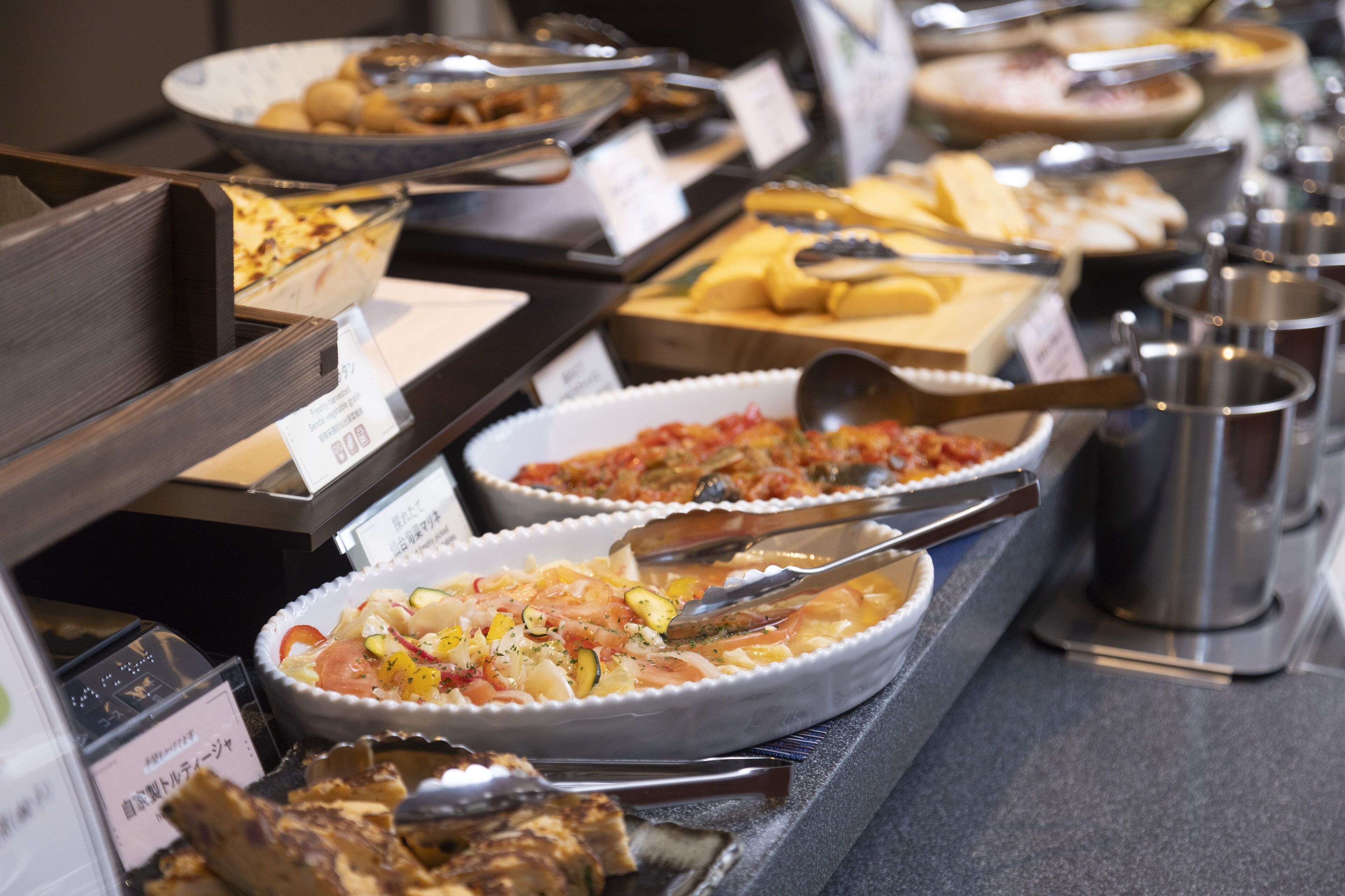 A buffet with various dishes, including pasta, salads, and bread, with serving utensils and labeled dishes.