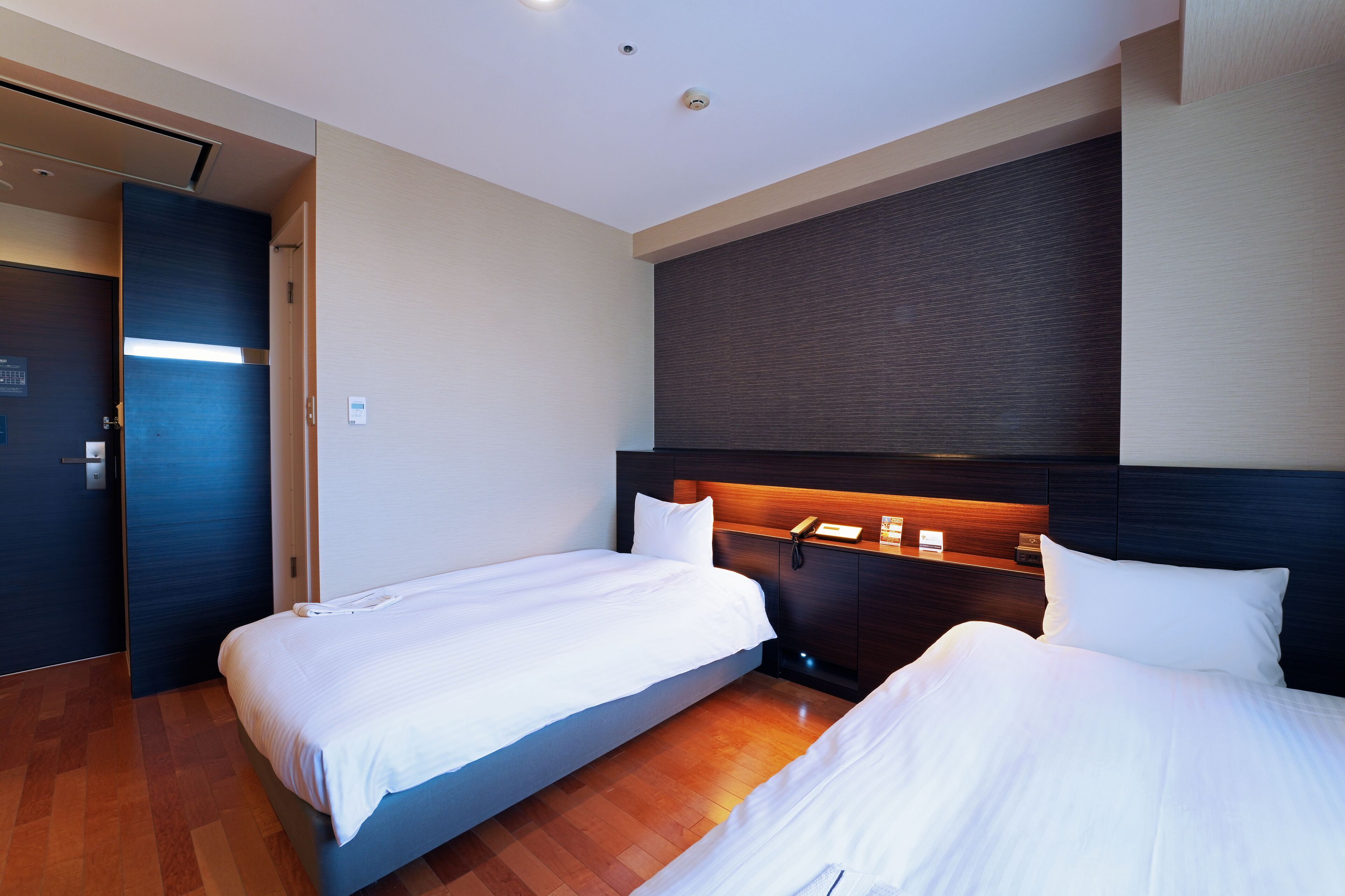 Ein Hotelzimmer mit zwei Einzelbetten, Holzboden, dunklem Kopfteil und einem kleinen Nachttisch mit Telefon und Lampe.