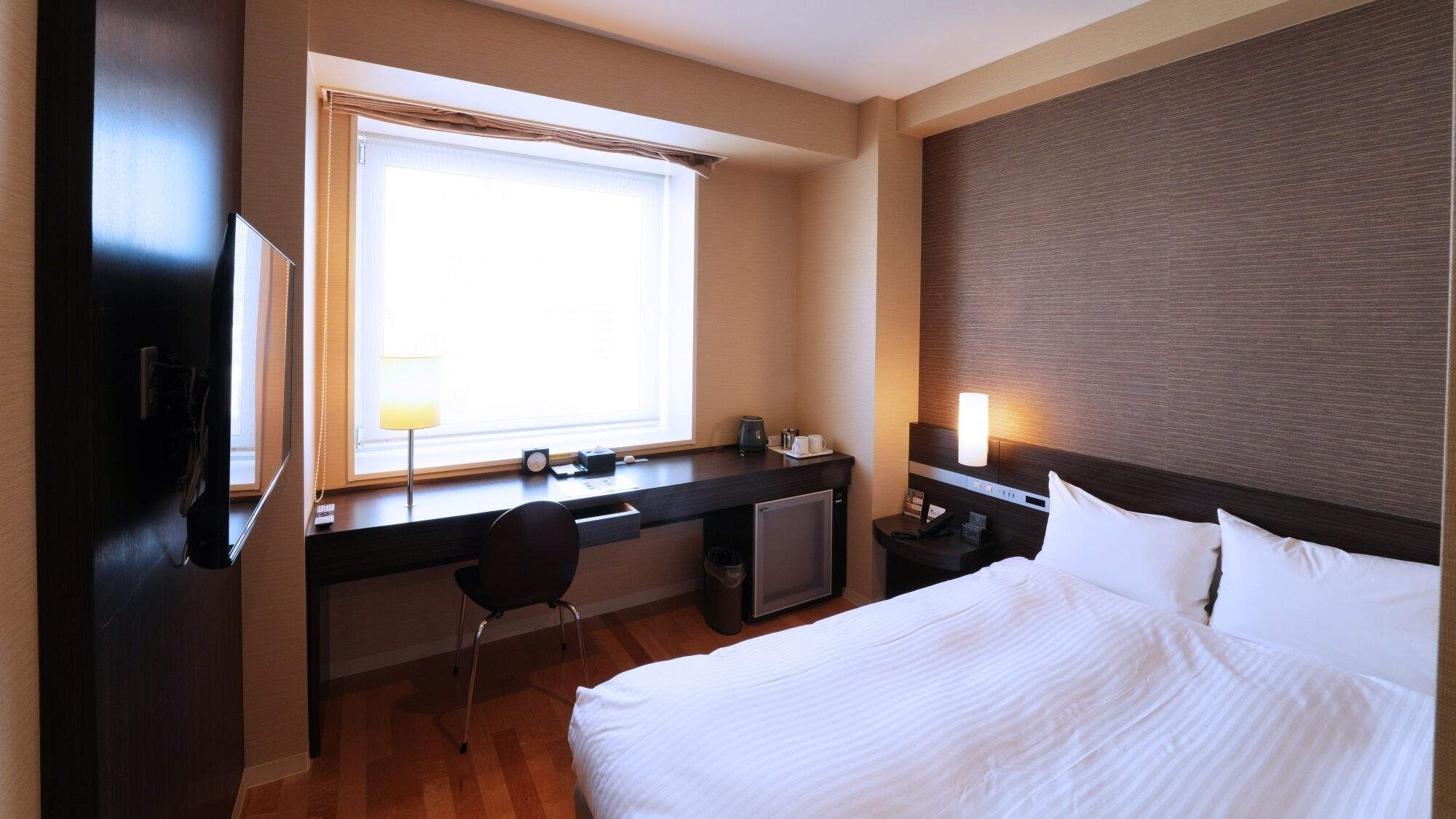 A hotel room with a bed, desk, chair, and a window with blinds.