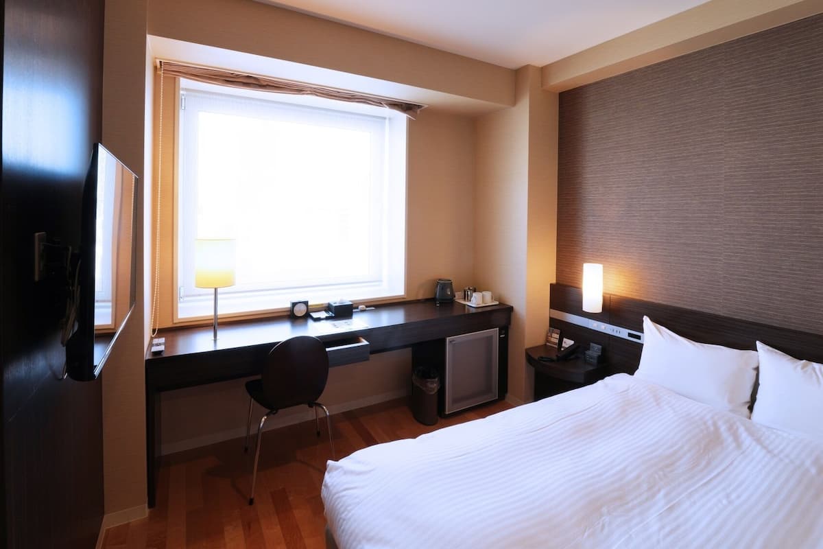 Quarto de hotel com cama, escrivaninha, cadeira e janela com persianas.