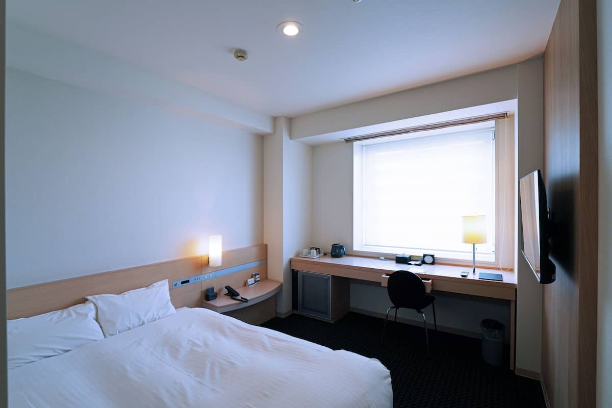 Quarto de hotel com cama, escrivaninha, cadeira e janela com persianas.