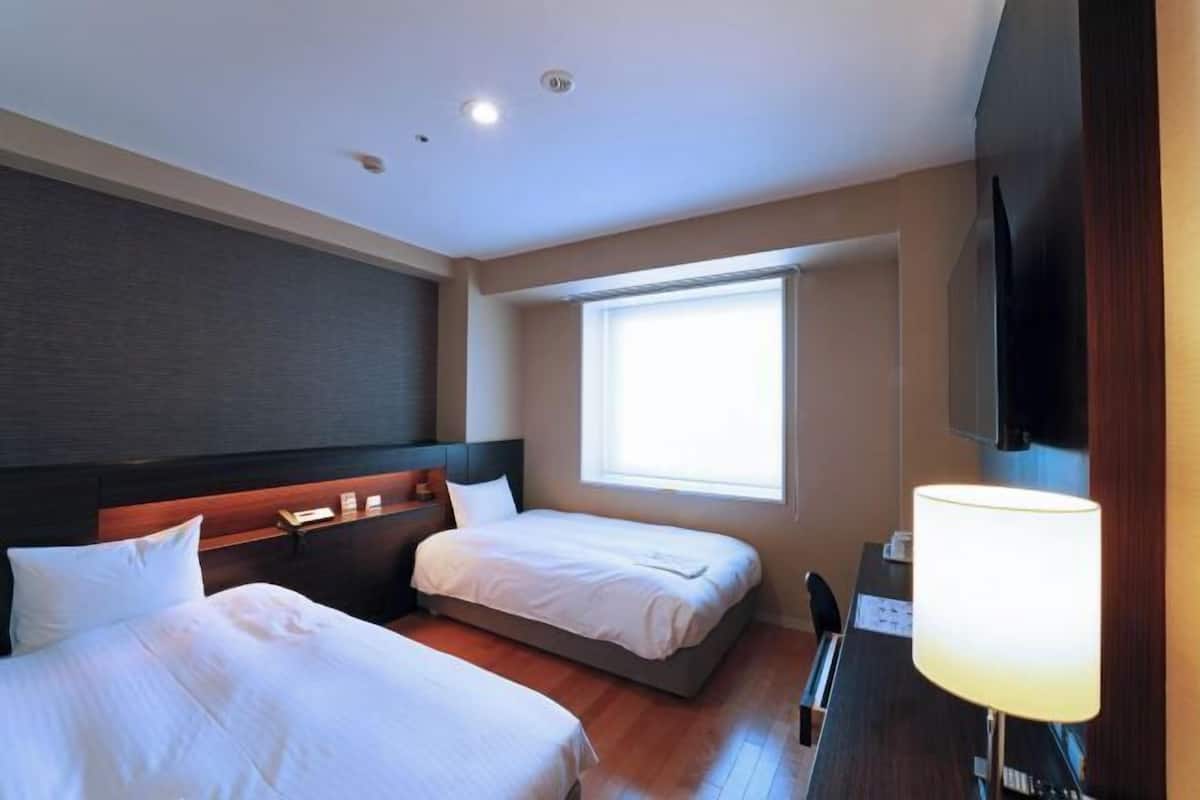 A hotel room with two beds, a desk with a lamp, a television, and a window.