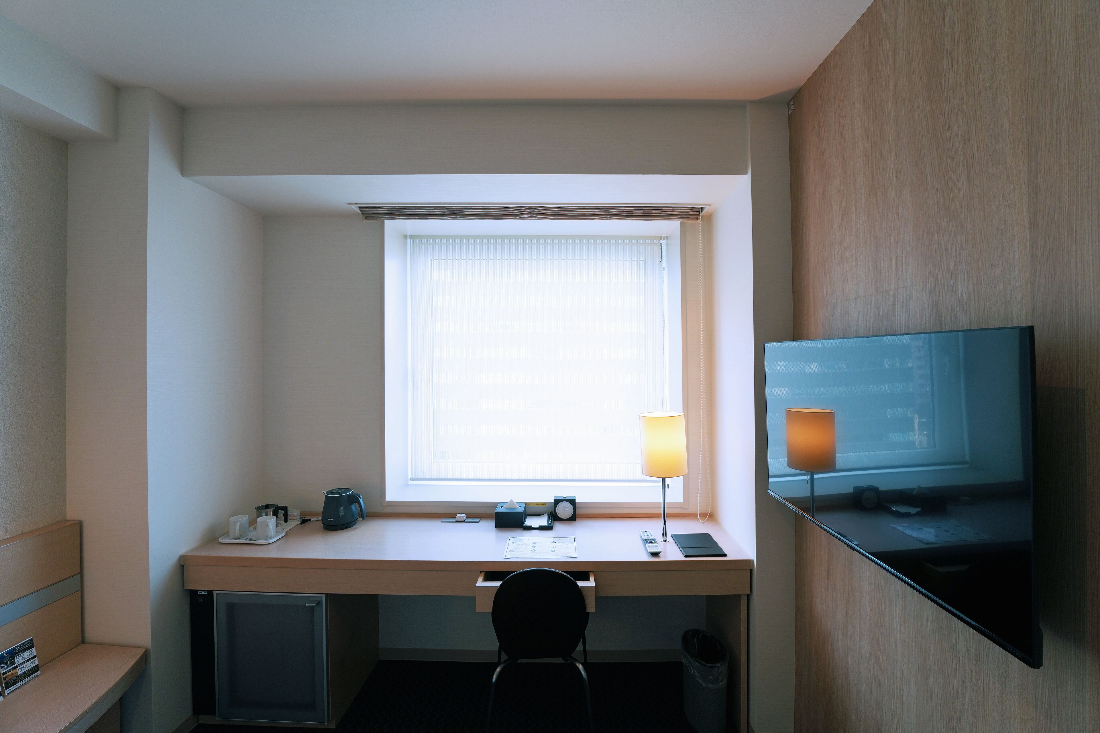 Une chambre d’hôtel équipée d’un bureau, d’une chaise, d’une télévision, d’une fenêtre avec des stores, d’une lampe et d’une poubelle.