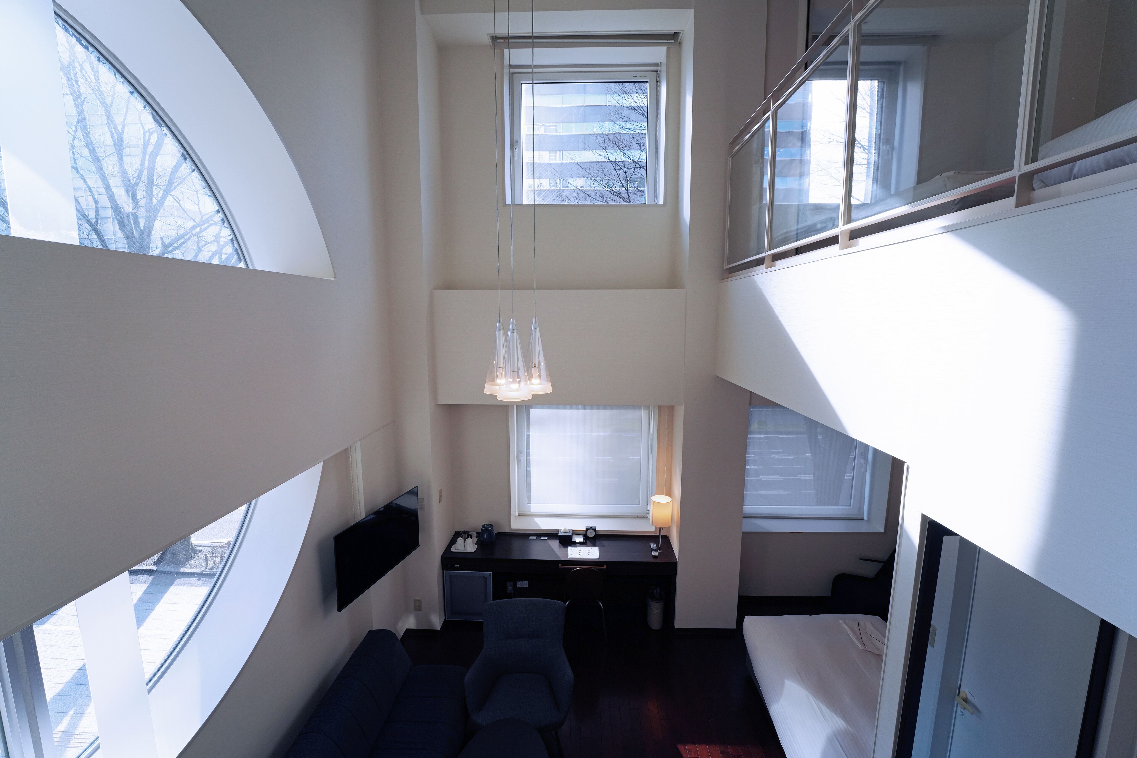 Una habitación de hotel moderna con un área de descanso en altura, un escritorio con lámpara y ventanas amplias que dejan entrar luz natural.