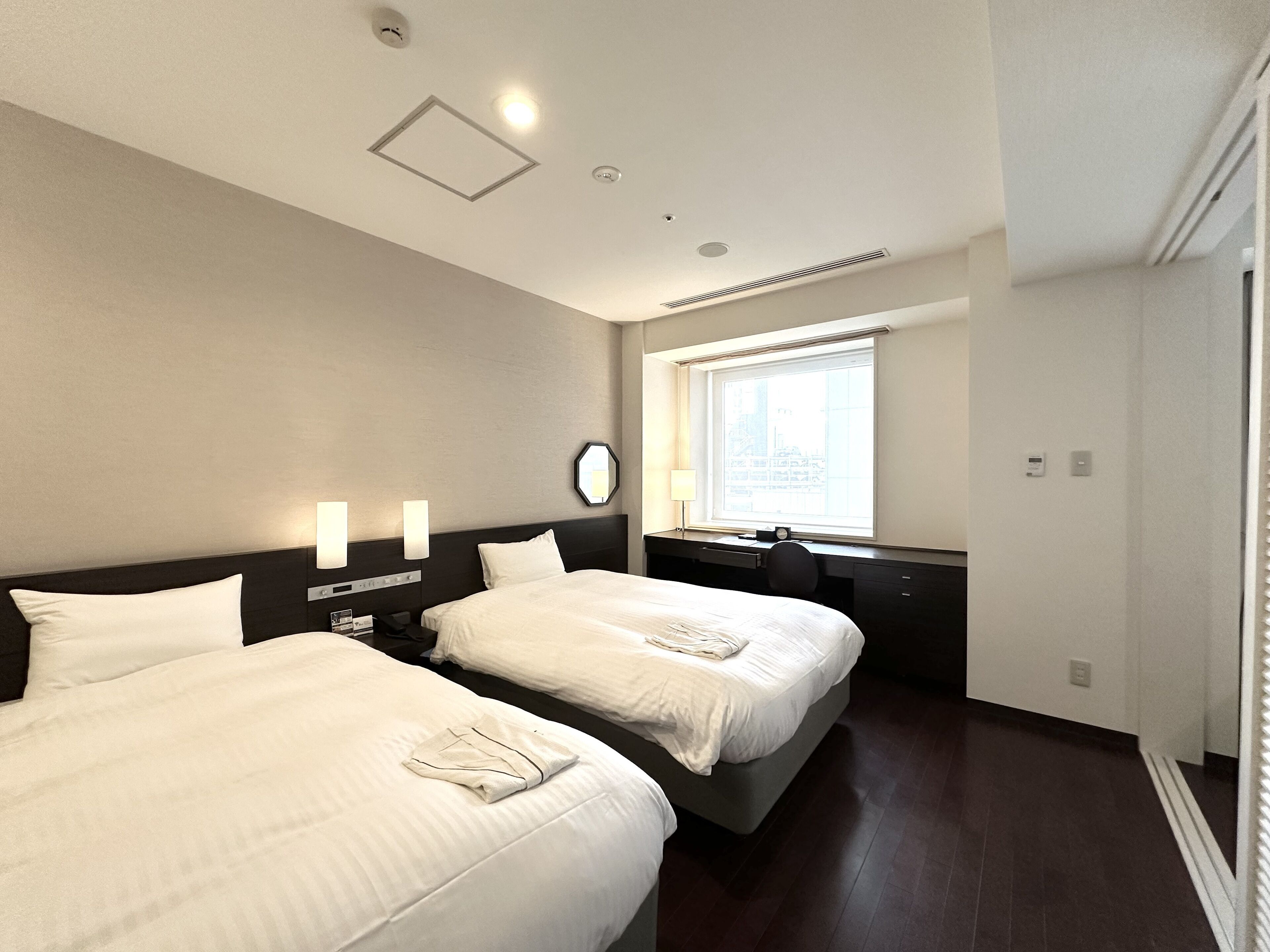 A hotel room with two beds, a desk, a chair, a window with a view, and a clock on the wall.