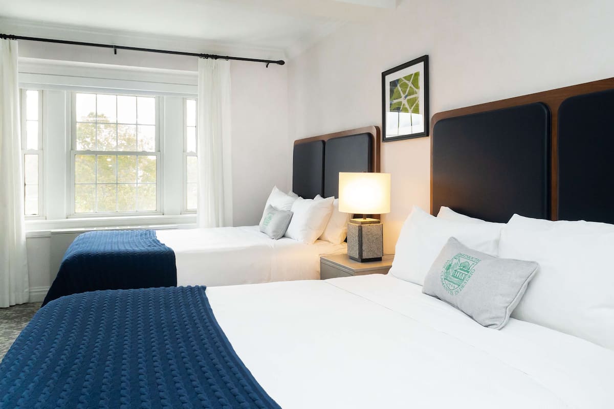 A hotel room with two beds, a window, a nightstand with a lamp, and a framed picture on the wall.