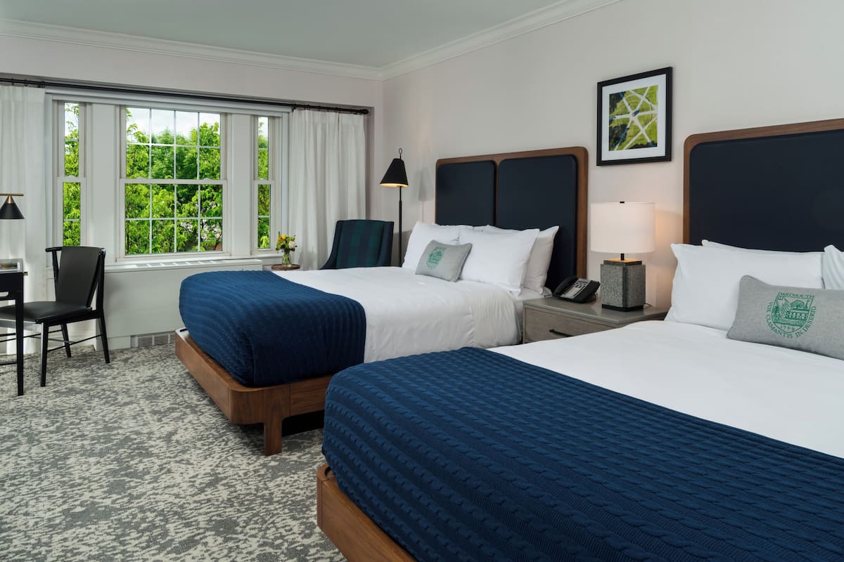A hotel room with two beds, a desk, a chair, a lamp, and a window with a view of trees.