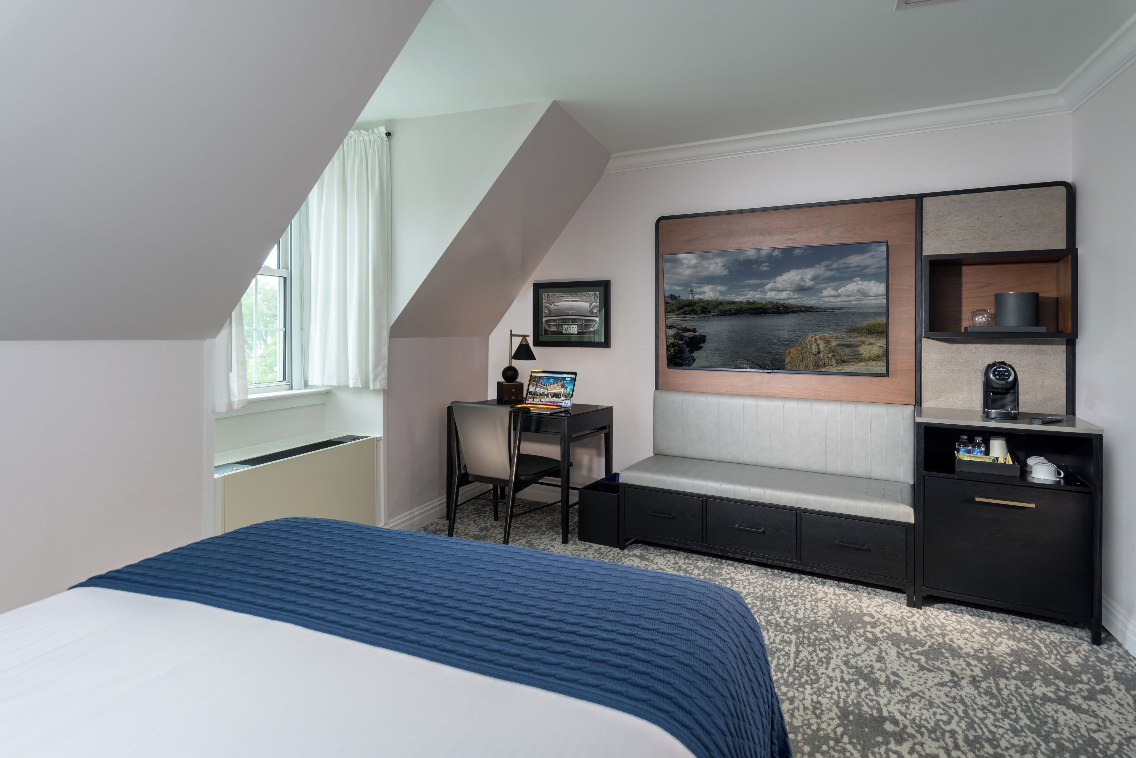 A hotel room with a bed, a desk, a chair, a bench, a large window, and a wall-mounted TV.