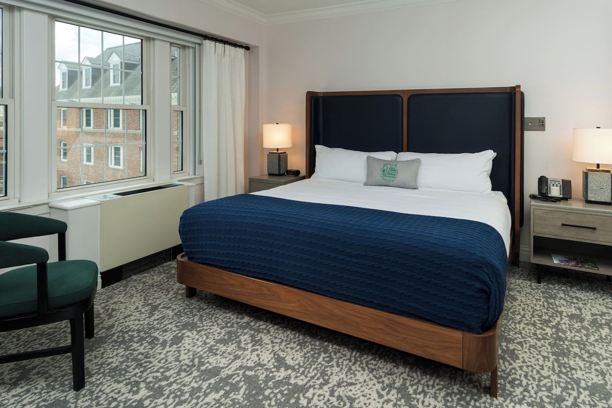 A hotel room with a large bed, a chair, a nightstand with a lamp, and a window with a view of buildings.