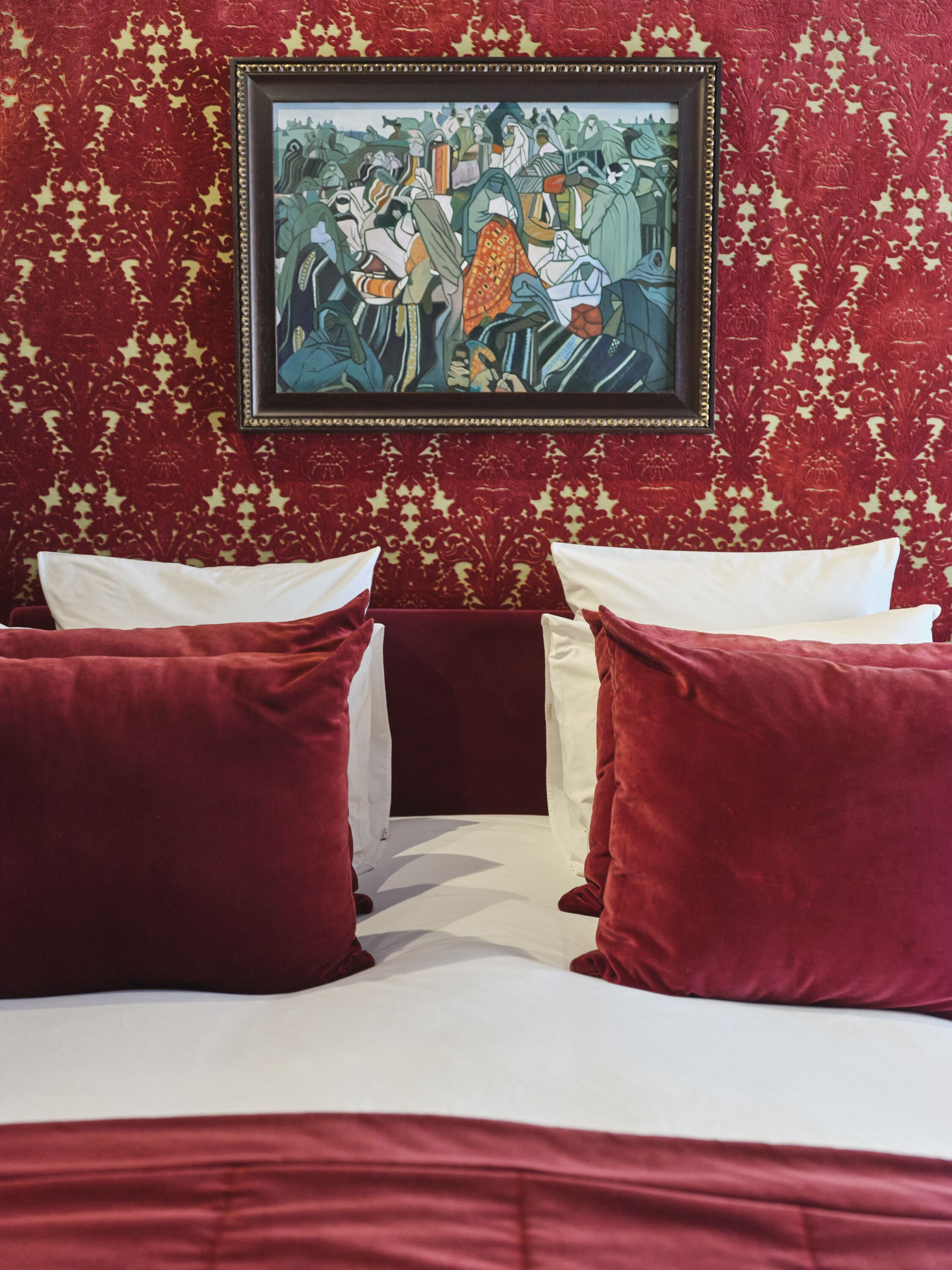 A hotel room with a bed, two red pillows, a framed artwork on the wall, and a patterned wallpaper.