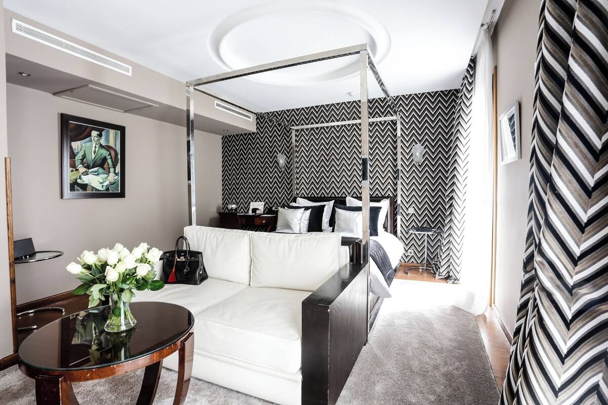 Ett modernt hotellrum med en säng med himmelssäng, en vit soffa, ett runt bord med en vas med blommor och en stor spegel.