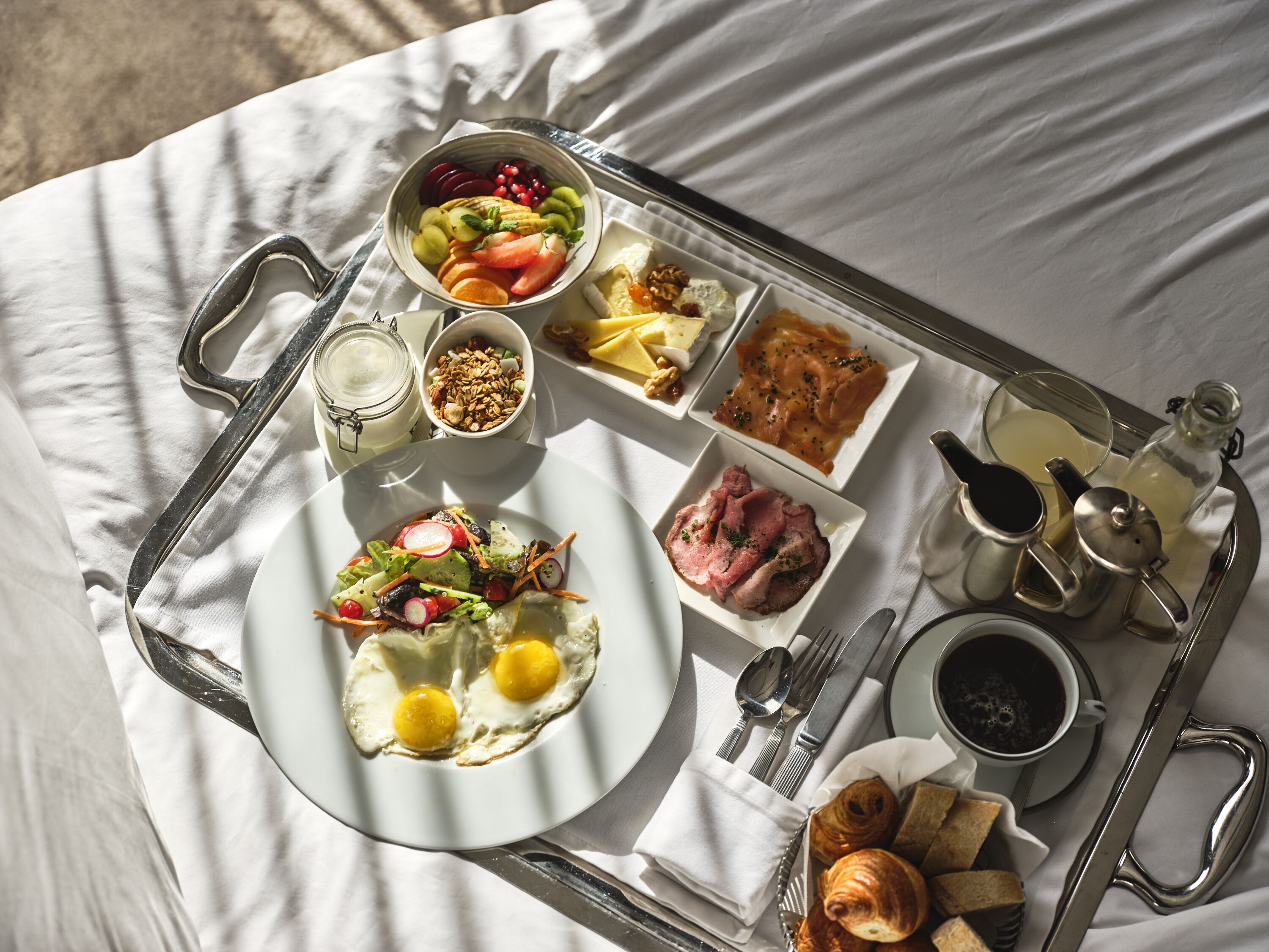 Bandeja de desayuno con huevos, fruta, jamón, cruasanes y café sobre un mantel blanco.