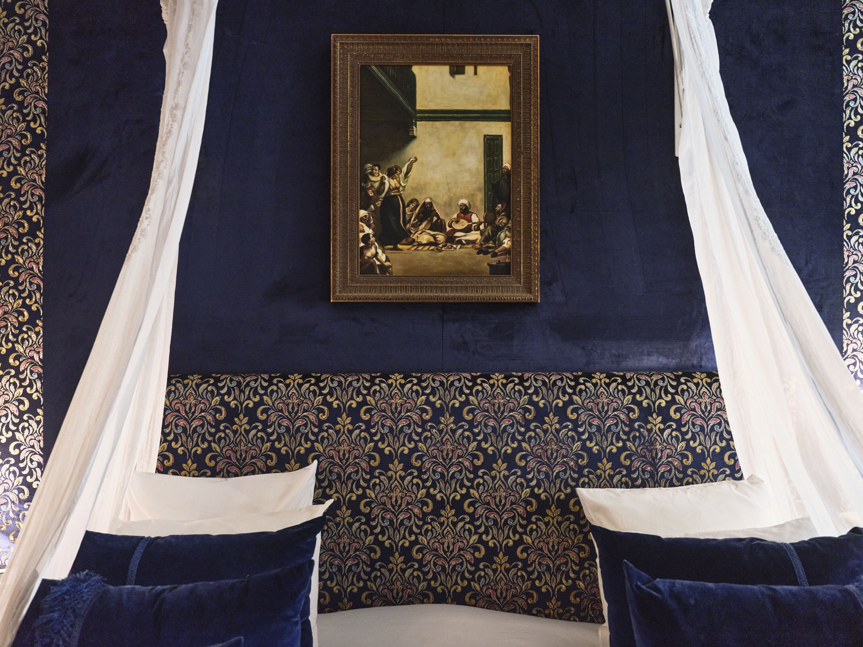 Une chambre à coucher avec un grand lit, un baldaquin, un papier peint à motifs et une œuvre d’art encadrée accrochée au mur.
