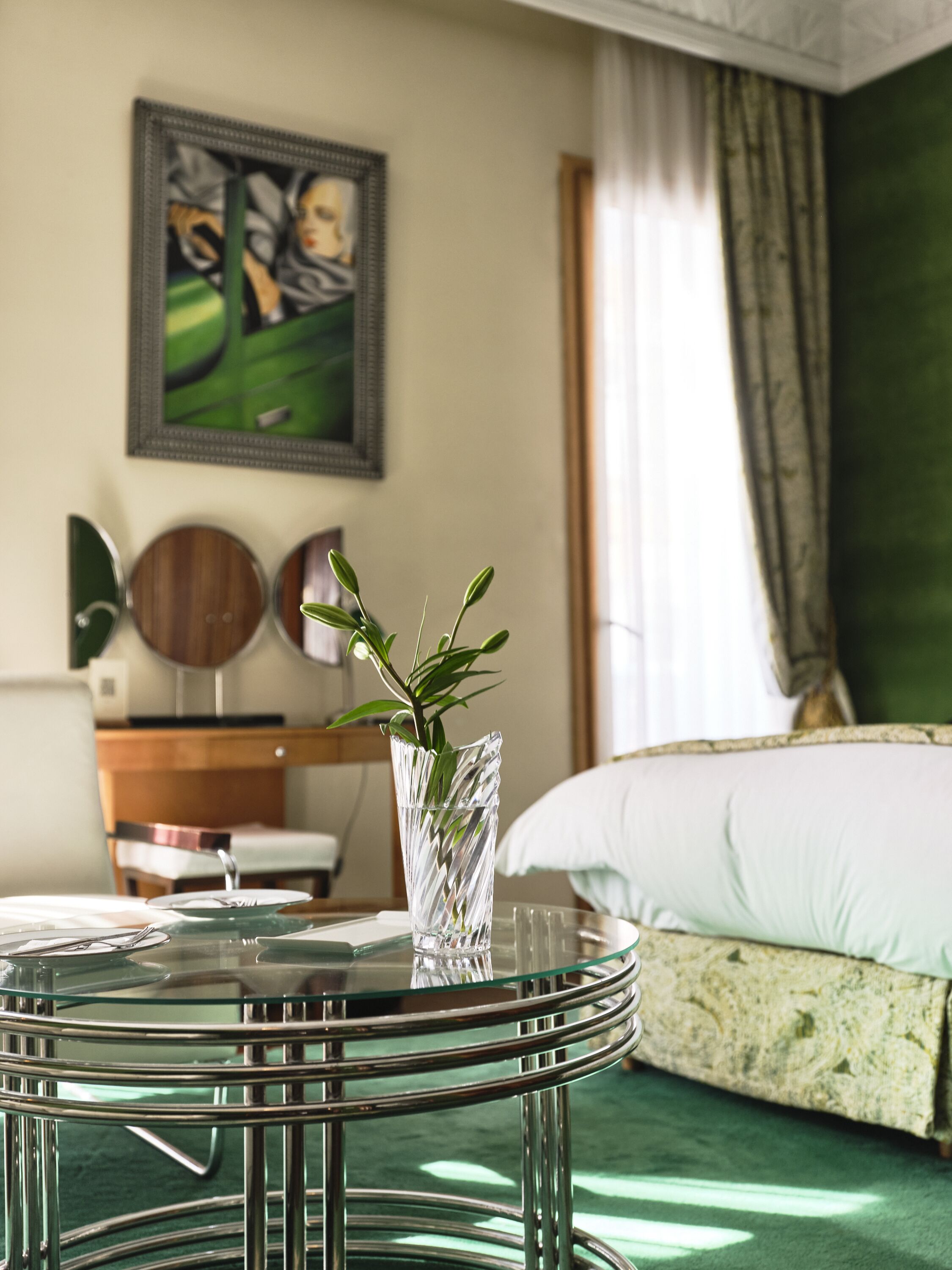 Una habitación de hotel con una cama, una mesita de vidrio con una planta, un cuadro en la pared y un espejo en la cómoda.