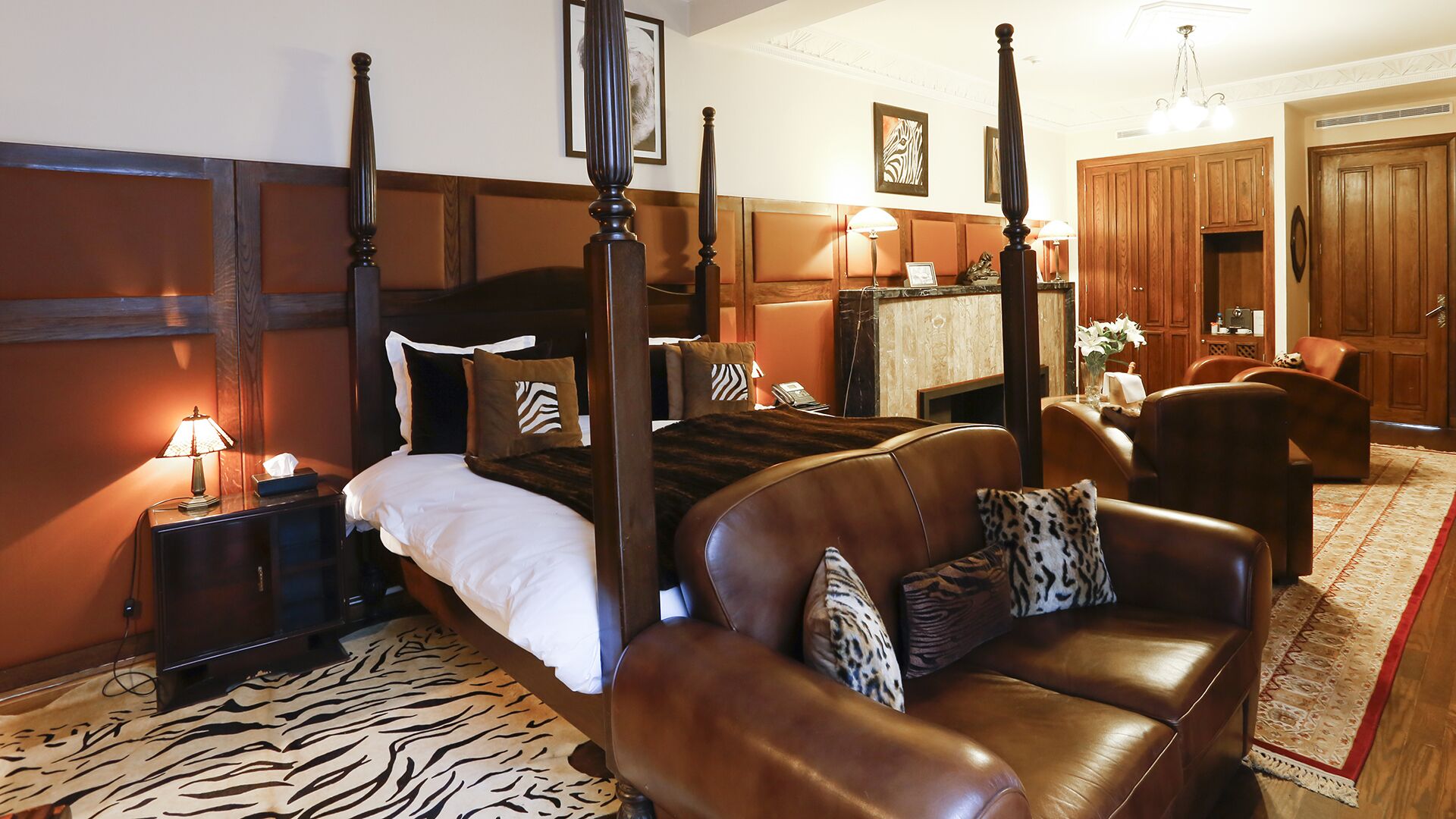 Una cama con dosel con un alfombra estampado de cebra, un sofá de cuero con cojines estampados, paneles de madera y un armario de madera en una habitación de hotel.