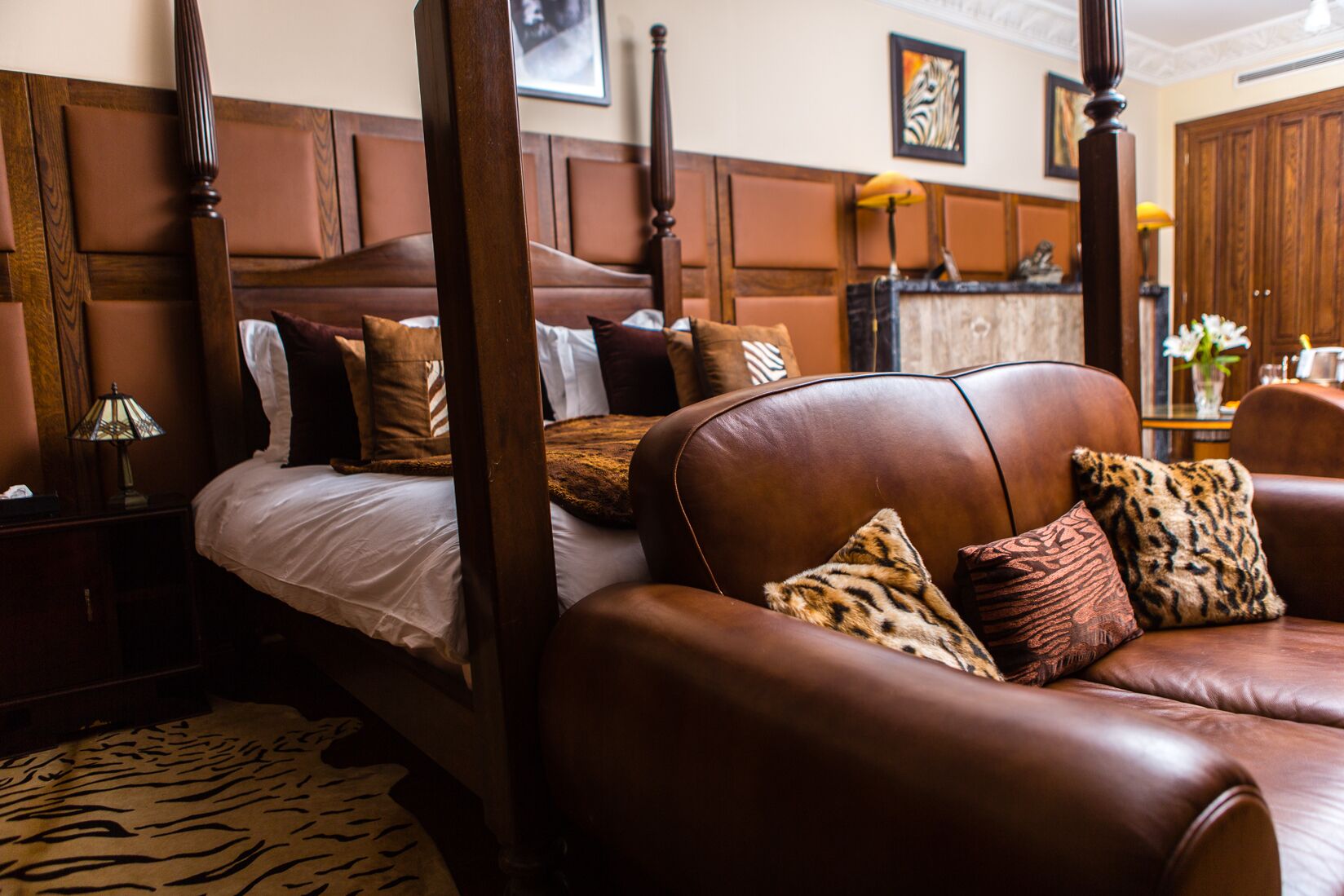A four-poster bed with a headboard, a leather sofa with patterned cushions, and a wooden nightstand with a lamp.