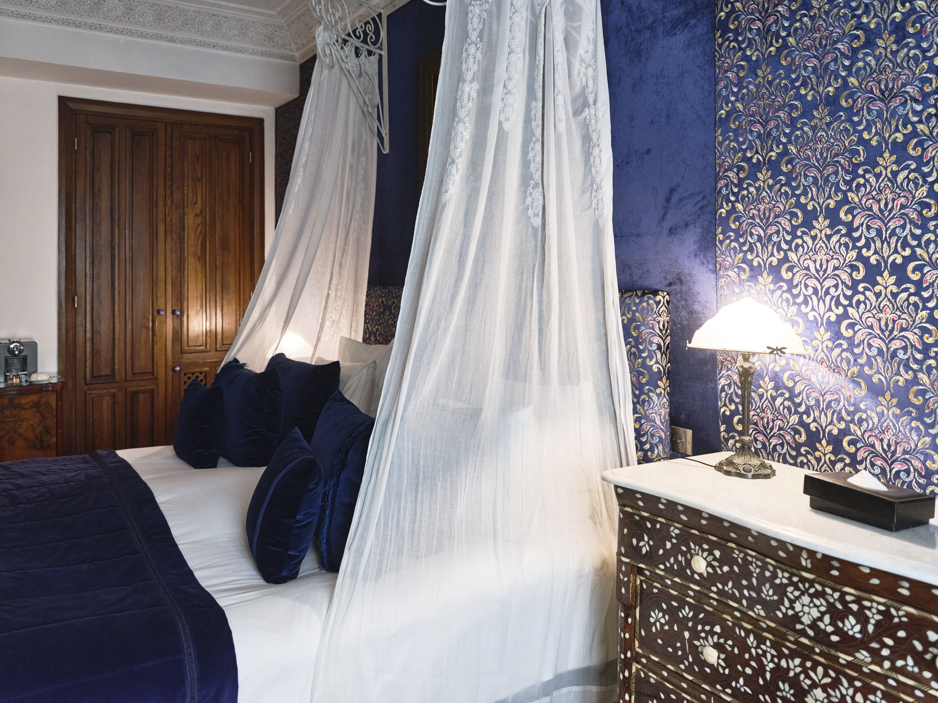 Un dormitorio con cama con dosel, una mesita de noche con lámpara y un papel tapiz estampado.