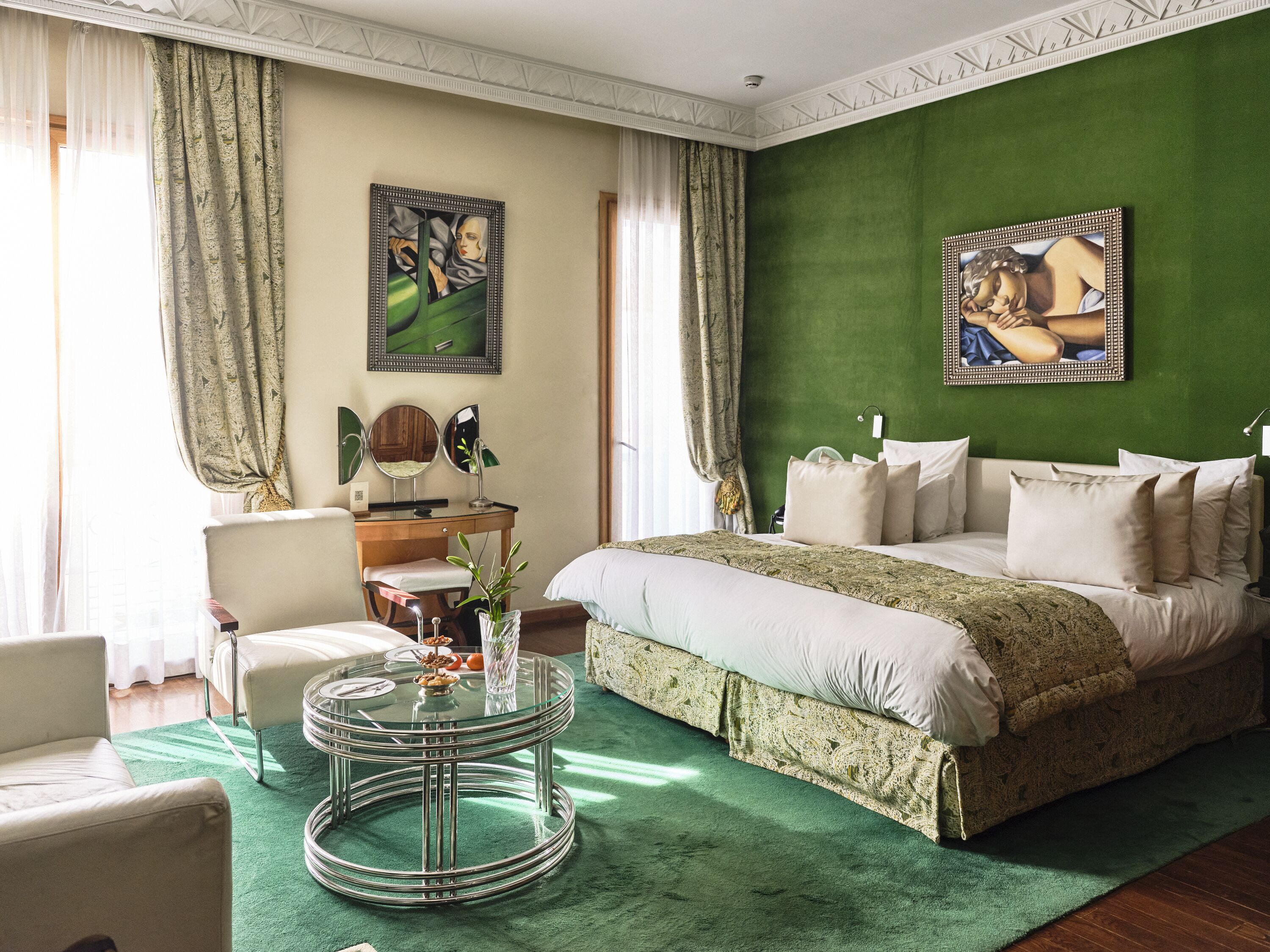 Un dormitorio con una pared de acento verde, una cama con muchos almohadones, una mesa redonda de cristal con un florero y un escritorio con un espejo.