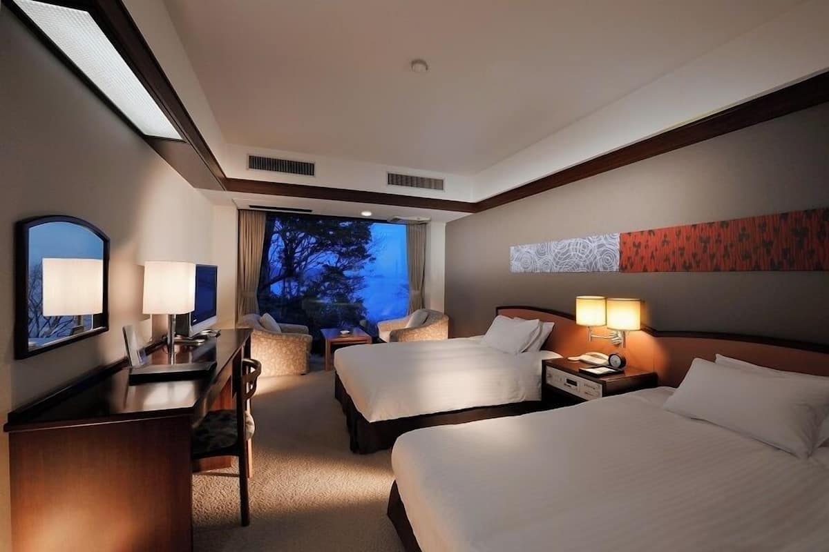 Habitación de hotel con dos camas, un escritorio, una silla, una mesita de noche y un ventanal con vistas a árboles.