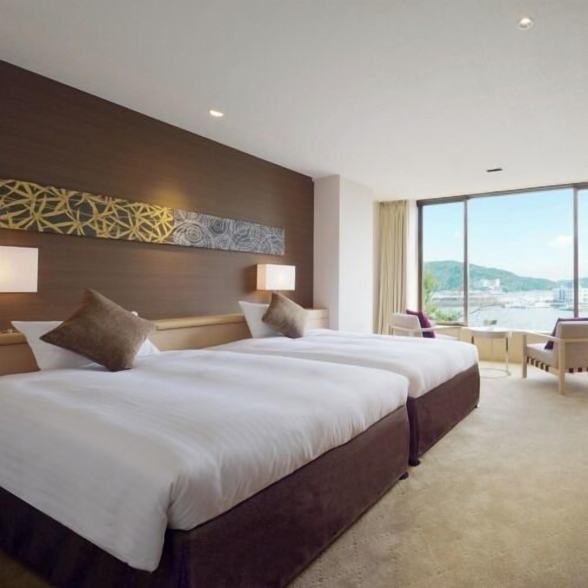 A hotel room with two beds, a large window with a city view, a desk, and a chair.