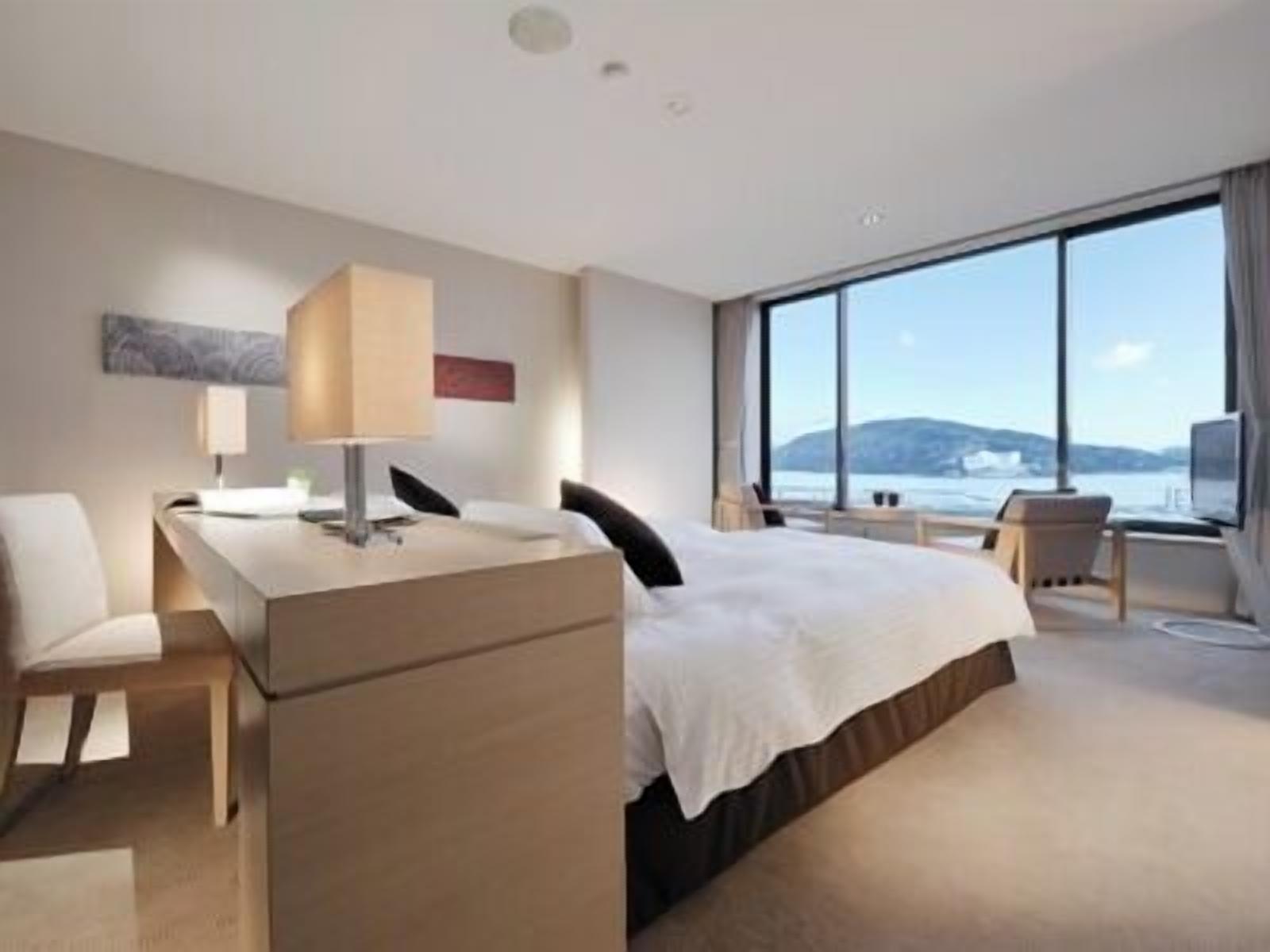 A modern hotel room with a bed, desk, chair, and large windows offering a view of the sea.