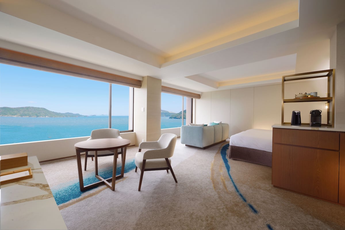 Habitación de hotel moderna con un amplio ventanal que ofrece vista al mar, un rincón de descanso acogedor, una pequeña mesa y un diseño minimalista.