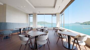 A modern dining area with tables and chairs, large windows with a view of the sea and hills.