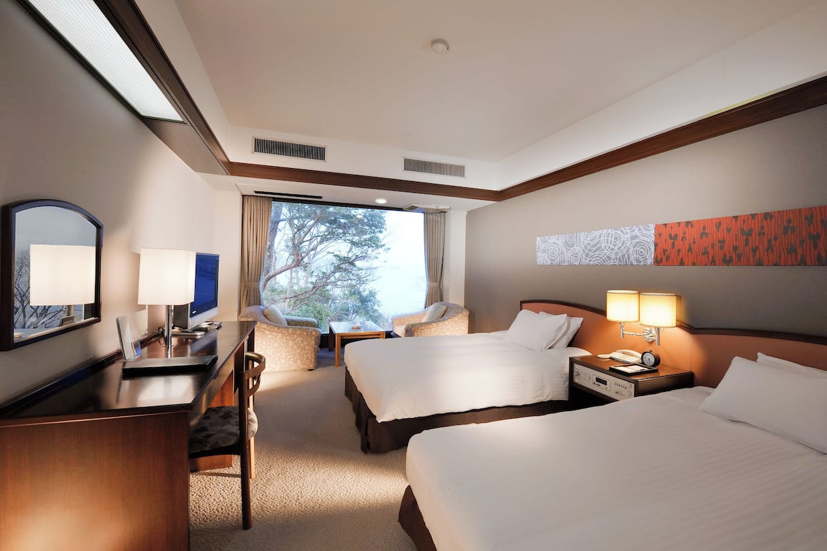 Habitación de hotel con dos camas, un escritorio con computadora, una silla y un ventanal con vistas a árboles.
