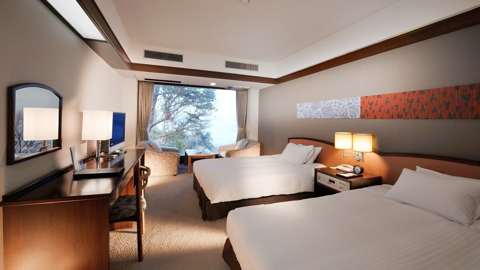 A hotel room with two beds, a desk, a chair, a TV, and a large window with a view of trees.