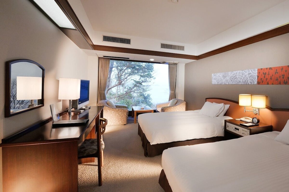 A hotel room with two beds, a desk, a chair, a TV, and a large window with a view of trees.
