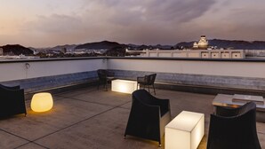 Una terraza en la azotea con mobiliario exterior, incluyendo sillas y una mesa, y vistas a un paisaje urbano y montañas al fondo.
